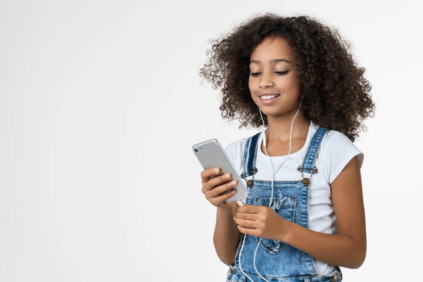 Happy african american girl smiling while using smartphone and earphones isolated over white background - Foto, Imagen