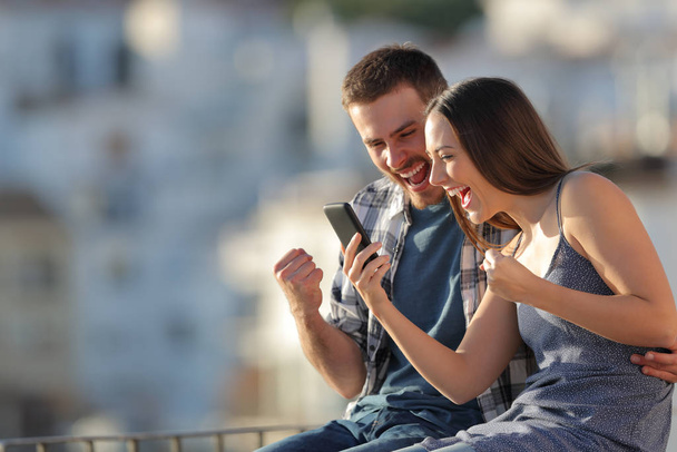 Excited couple celebrating online smart phone news outdoors in a town outskirts at sunset - Photo, image