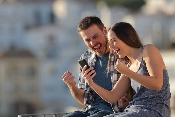 Excited couple or friends reading smart phone content in a town at sunset - Foto, afbeelding