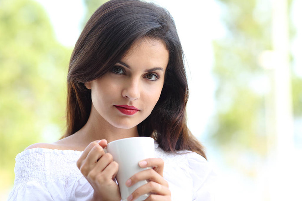 Front view portrait of a sexy woman holding a cup of coffee looking at camera outdoors - Фото, зображення