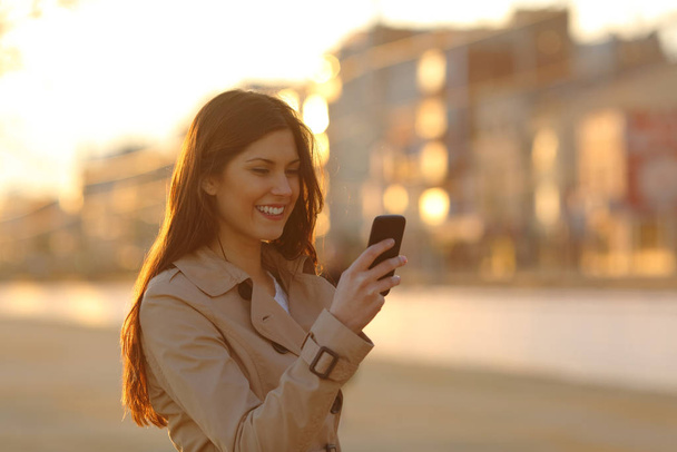 Happy woman using a smart phone walking at sunset in the street - Valokuva, kuva