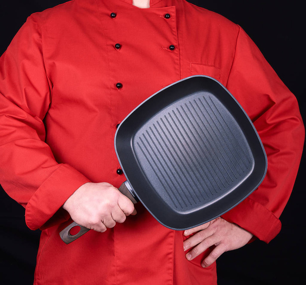 cook in red uniform holding an empty square black frying pan, black background - Fotoğraf, Görsel