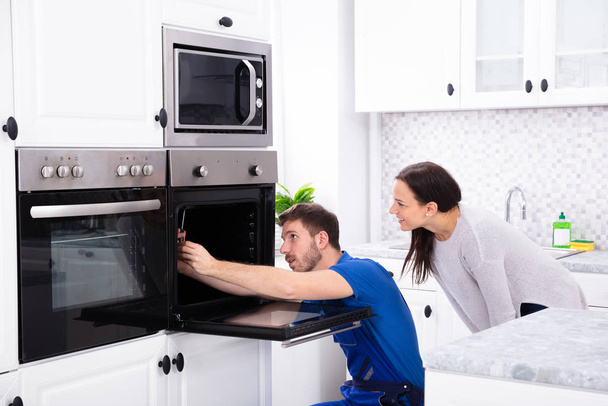 Smiling Young Woman Looking At Technician Fixing Oven In Kitchen - Photo, image