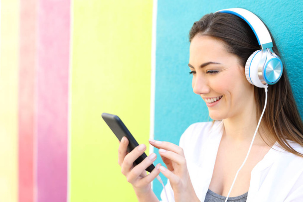 Happy woman listens to music from smart phone leaning on a wall in a colorful street - Photo, image
