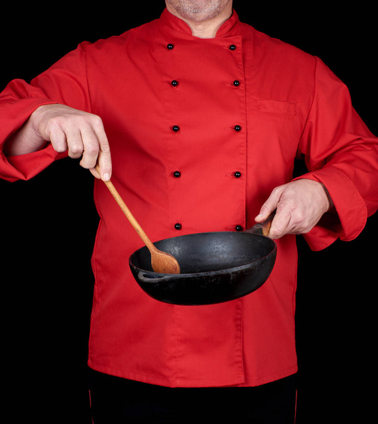 cook in red uniform holding an empty  black frying pan and wooden spoon, black background - Foto, afbeelding