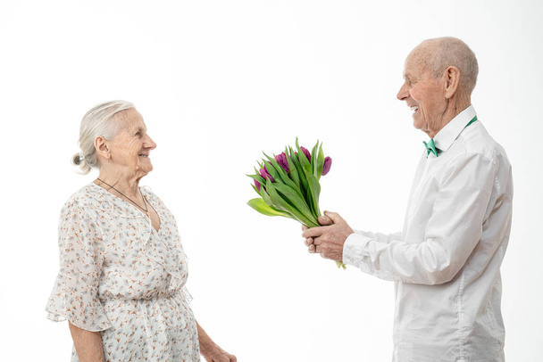 Senior homme en chemise blanche tenant des fleurs dans les mains sur un rendez-vous avec une femme âgée en robe blanche isolée sur fond blanc, vieux mari donne un bouquet de tulipes à sa femme
 - Photo, image
