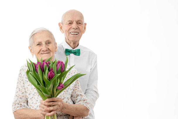 Waist up portrait of the happy smiling old couple, senior in white shirt hugging his wife in white dress that holds flowers in hands, both are looking at the camera isolated over white background - Photo, Image