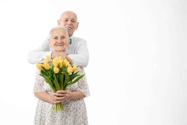 Feliz pareja de ancianos sonrientes en ropa blanca, la mujer sostiene flores amarillas en las manos de su marido mayor se para detrás y mirando a la cámara aislada sobre el fondo blanco, copyspace para su texto
 - Foto, Imagen
