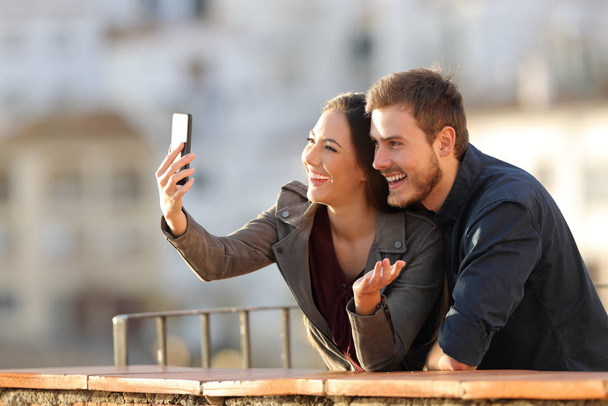 Happy couple having a video call or taking selfies with a smart phone in a balcony at sunset - Φωτογραφία, εικόνα