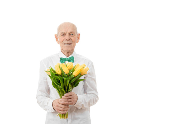 Szczęśliwy, zadowolony, wesoły, radosny uśmiechnięty starzec w białej koszuli, senior trzyma bukiet żółtych kwiatów w rękach i patrząc na aparat odizolowany na białym tle, copyspace dla Twojego tekstu - Kobiety - Zdjęcie, obraz