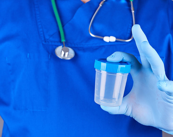 the doctor in blue uniform and latex gloves is holding an empty plastic container for taking urine samples, close up - Foto, Imagem