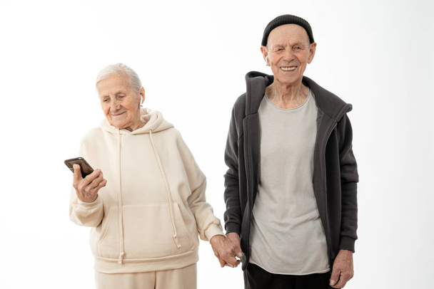 Allegro anziano elegante in felpa con cappuccio e cappello nero tiene la mano di sua moglie che guardando il suo telefono, isolato su sfondo bianco
 - Foto, immagini