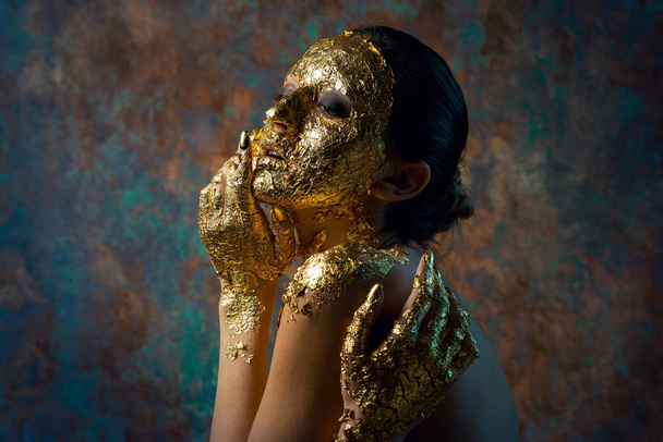 Tyttö, jolla on kultaisesta lehdestä tehty naamio kasvoillaan. Gloomy studio muotokuva brunette abstrakti tausta. - Valokuva, kuva
