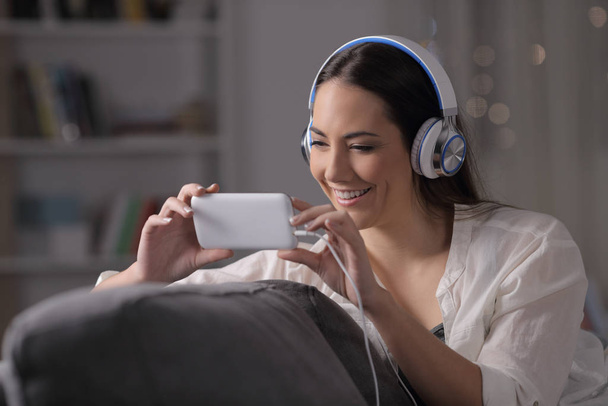 Mulher feliz assiste vídeos usando fones de ouvido sentados em um sofá à noite em casa
 - Foto, Imagem