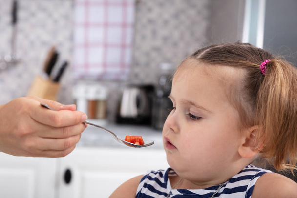 Close-up Of Mother's Hand Feeding Tomato Slice To Her Daughter With Spoon In Kitchen - Zdjęcie, obraz
