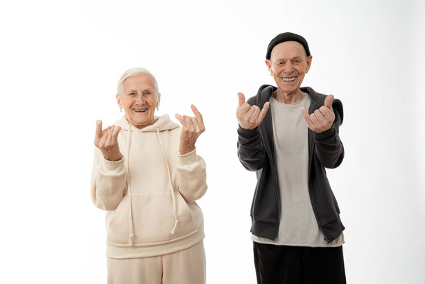 Привлекательные пожилые люди пара в толстовках, старик и женщина делают рок-н-ролл жест и глядя на камеру изолированы на белом фоне
 - Фото, изображение
