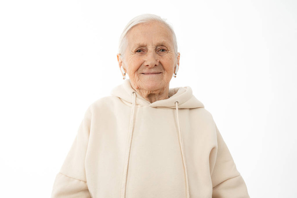 Close up photo of attractive old woman in beige hoodie with white airpods in ears looking at the camera isolated over white background - 写真・画像