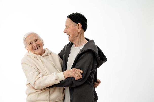 Gelukkig glimlachen positieve pencioner paar in hoodies knuffelen, stijlvolle oude man en vrouw geïsoleerd over witte achtergrond, copyspace voor uw tekst - Foto, afbeelding
