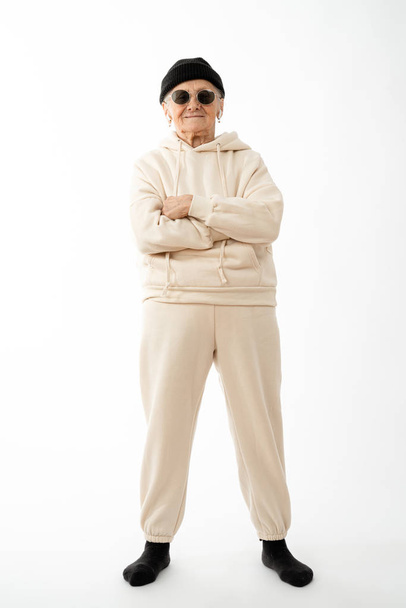 Stylish old hipster woman in beige hoodie black hat and sunglasses stands with crossed hands and looking at the camera isolated over white background - 写真・画像