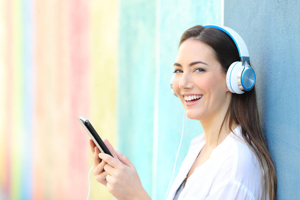 Happy woman looking at you listening to music from tablet in a colorful street - Фото, изображение