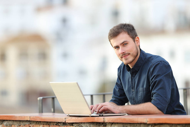 Confident man using a laptop and looking at camera in a balcony in a town - Foto, imagen