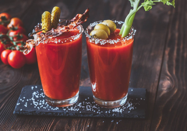 Two glasses of Bloody Mary garnished with gherkins, fried bacon strips, green olives and celery stalk - Fotoğraf, Görsel