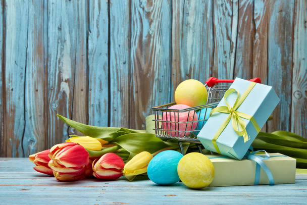 Colored easter eggs in shopping trolley, gift boxes and spring tulips flowers on a wooden  background with copy space. Easter background, Easter shopping. - Фото, зображення