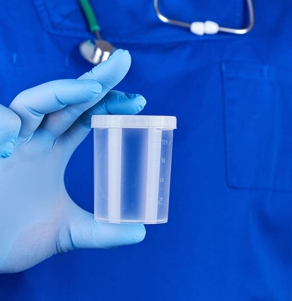 doctor in blue uniform and latex gloves is holding an empty plastic container for taking urine samples, close up - Foto, Imagem