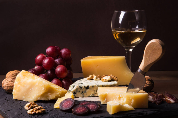 Assorted cheeses, nuts, grapes, fruits, smoked meat and a glass of wine on a serving table. Dark and Moody style. Free space for text. - Zdjęcie, obraz