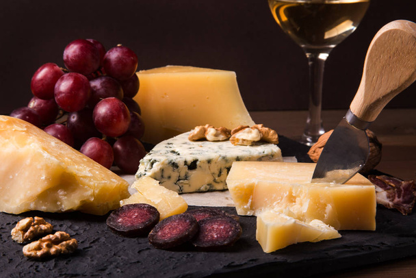 Assorted cheeses, nuts, grapes, fruits, smoked meat and a glass of wine on a serving table. Dark and Moody style. Free space for text. - Photo, image