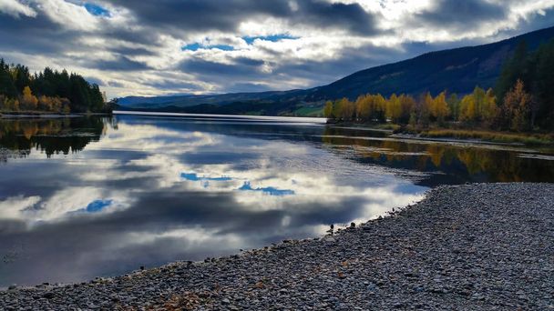 Peaceful and quiet untouched landscape scenery of pebble beach and lake with dramatic clouds reflected in the calm water with autumn trees around and rural   mountains in the horizon. - Fotografie, Obrázek