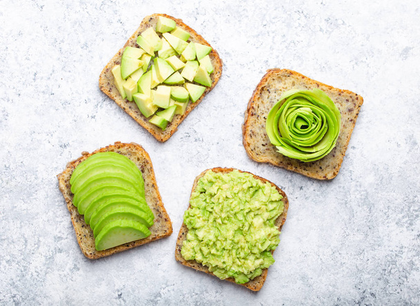 Concept of healthy breakfast toasts with avocado smashed, slices, chunks and rose. Different ways of making and serving delicious avocado sandwich, top view, white stone background, close-up - Фото, зображення