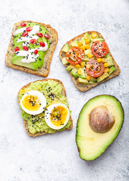 Avocado toasts with egg, tomatoes, seasonings and a half of whole avocado over white stone background. Healthy breakfast avocado sandwiches with different toppings, top view, close-up. - Foto, Imagem