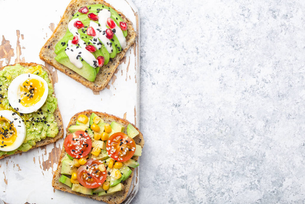 Avocado toasts with egg, tomato, seasonings on white wooden rustic cutting board, stone background. Making healthy breakfast avocado sandwiches, different toppings, top view, close-up, space for text. - Foto, imagen