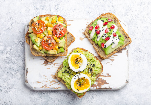 Avocado toasts with egg, tomatoes, seasonings over white wooden rustic cutting board on stone background. Making healthy breakfast avocado sandwiches with different toppings, top view, close-up. - Foto, Imagem