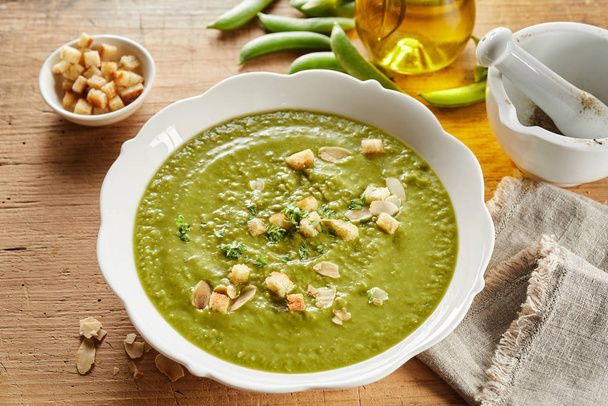 Bowl of thick pea soup served with sauteed croutons, flaked almonds and sprinkled with herbs viewed hogh angle with fresh ingredients and olive oil - Foto, afbeelding