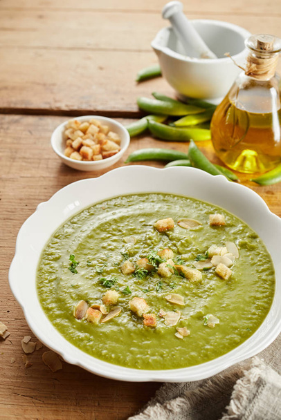 Homemade pea soup with roasted almonds and crunchy toasted bread croutons garnished with herbs viewed high angle on a rustic table - Photo, image