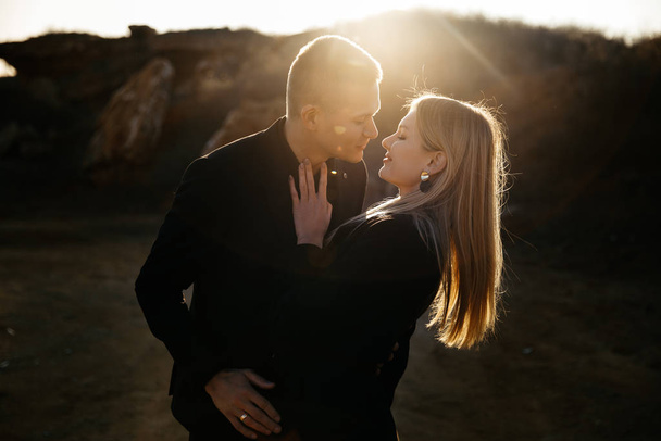 Retrato vista lateral de dos jóvenes amantes besándose al atardecer sobre fondo barranco. Pareja en ropa negra
 - Foto, imagen