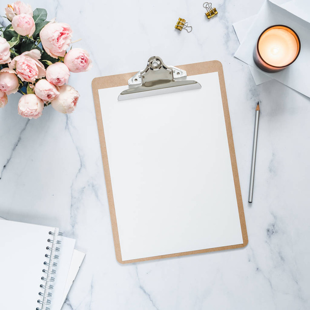 Top view of clipboard with white empty page. Clipboard, flowers, scented candle on white marble. Feminine home office mock up with blank sheet of paper A4 portrait format,copy space for text. Flat lay - Фото, изображение