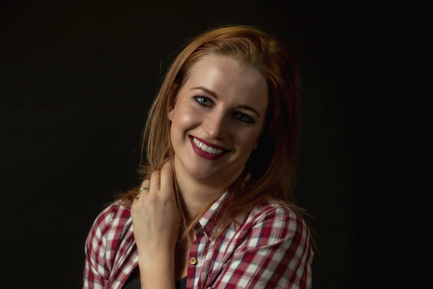 Low key portrait of smiling young woman dressed in a plaid shirt and holding her neck by hand. - Фото, изображение