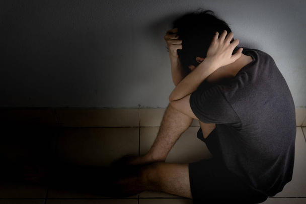 θλιμμένος άντρας αγκαλιάζει το γόνατό του και κλαίει καθισμένος μόνος σε ένα σκοτεινό δωμάτιο. Στέρηση - Φωτογραφία, εικόνα