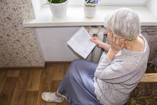 The senior woman holding gas bill in front of heating radiator. Payment for heating in winter. - Фото, изображение