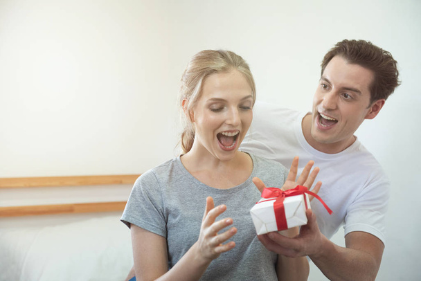 Felice giovane coppia caucasica che festeggia a casa. Bello fidanzato uomo dà la sua ragazza con scatola regalo sorprendentemente
. - Foto, immagini
