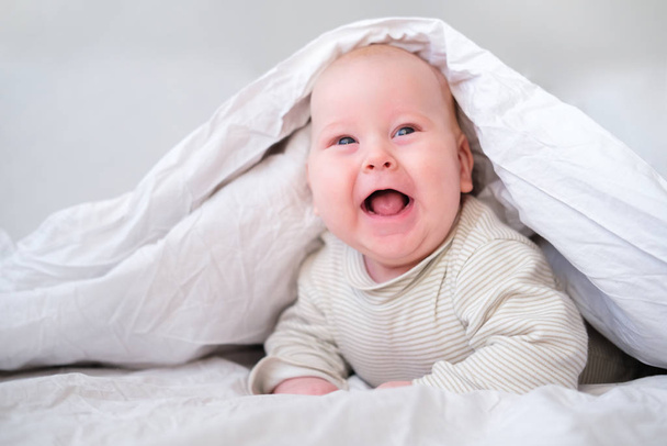 Adorable smiling baby boy under white blanket in bedroom. Newborn child relaxing in bed. Family morning at home. New born infant kids - Foto, Imagem