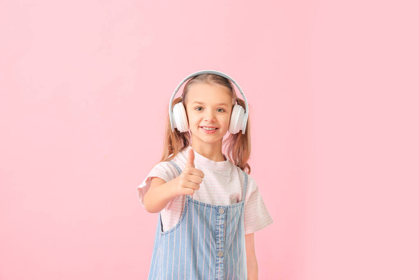 Cute little girl with headphones showing thumb-up gesture on color background - Foto, afbeelding