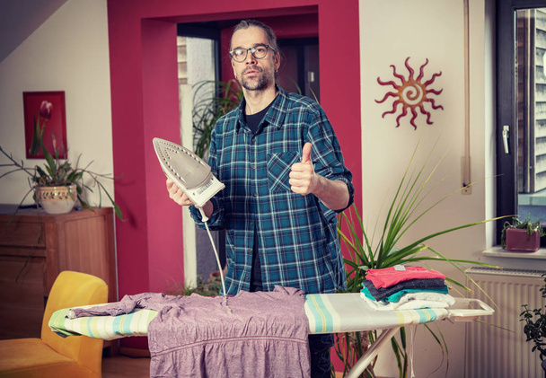 man with iron on ironing board with thumb up in living room - Photo, Image