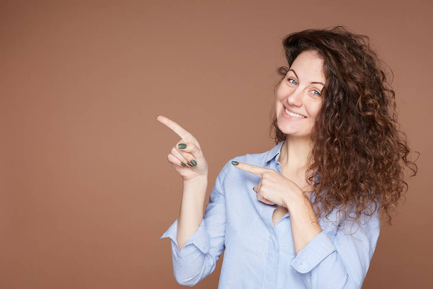 Curly headed lovely woman with pleasant look shows blank copy space for advertisement or information, has cheerful face expression, wears blue jacket, suggests to select option over pastel beige wall. - Foto, imagen