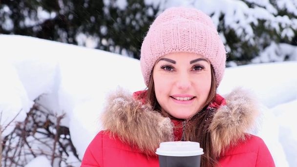 Jovencita feliz con una bebida caliente en un parque de invierno. Mujer en una chaqueta roja bebe café para llevar en un día soleado de invierno, nevando
 - Foto, imagen