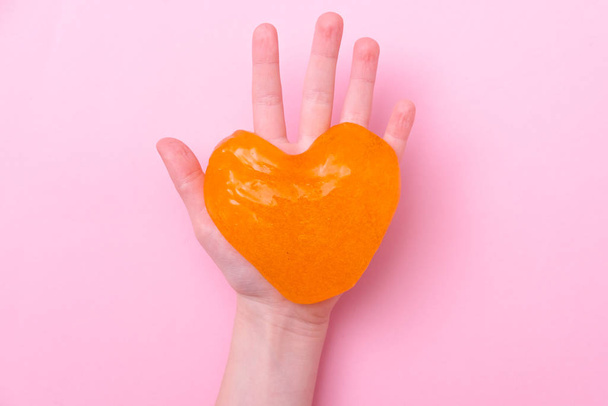 Orange slime in heart shape in kid hands. Girl hands playing slime toy on pink background. Making slime. Love and valentines day concept - Foto, imagen