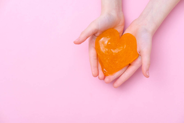Orange slime in heart shape in kid hands. Girl hands playing slime toy on pink background. Making slime. Copyspace. Love and valentines day concept - Foto, afbeelding